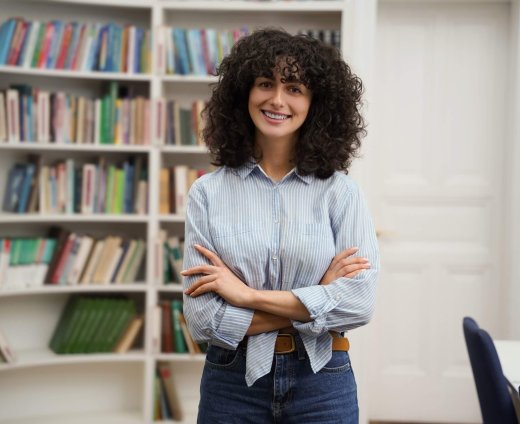 Curly-haired,Pretty,Young,Woman,Standing,On,The,Bookshelves,Background